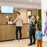 Twee families melden zich aan bij de receptiebalie van het Auris Audiologisch Centrum. Achter de balie staan twee medewerkers. Op een scherm is het Auris-intranet zichtbaar.
