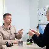 Een man overlegt met een audioloog. Op tafel liggen hoortoestellen en een doosje. De audioloog houdt een hoortoestel vast en legt iets uit.