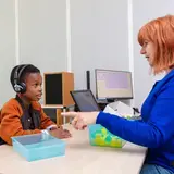 Een kind met grote koptelefoon doet een gehoortest. Tegenover hem zit een audioloog die de test begeleidt. Op tafel staan bakjes met gekleurde blokjes.