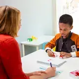 Een kind werkt met rode blokjes aan een taak. Tegenover hem zit een therapeut die aantekeningen maakt. Het kind is geconcentreerd bezig.