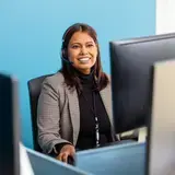 Een medewerker van het Auris Audiologisch Centrum zit aan haar bureau met een headset op. Ze glimlacht in de camera. Op de achtergrond is een blauwe muur zichtbaar.