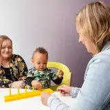 Een baby speelt met een geel speelgoedrek aan een tafel. Naast het kind zit de moeder. Tegenover hen zit een therapeut die het kind begeleidt.