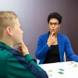 Een logopedist laat een jongen zien hoe hij zijn lippen moet bewegen. Ze wijst naar haar mond. Op tafel liggen kaartjes. De jongen kijkt haar na.