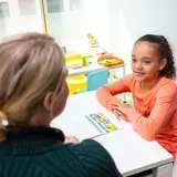 Een meisje zit tegenover een therapeut en kijkt haar glimlachend aan. Op tafel ligt een ringband open. Op de achtergrond zijn tafeltjes met speelgoed zichtbaar.