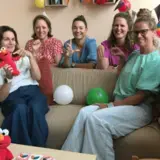 Vijf vrouwen zitten op een bank met een Elmo-knuffel en ballonnen, tijdens een opening.