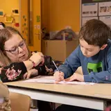 Een leerkracht helpt een leerling met schrijven aan tafel.