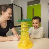 Een leerling doet een logopedie-oefening met een geel knutselwerk. De logopedist kijkt lachend toe.
