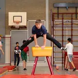 Een jongen maakt een spreidsprong over de pegasus in de gymzaal, terwijl een leraar en klasgenoten op de achtergrond toekijken.
