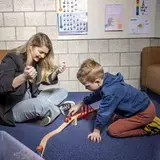 Een logopedist en een jongen spelen samen met een houten treinbaan en brug op de grond.