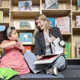 Een logopedist leest voor uit een prentenboek aan een meisje, tussen boekenkasten.