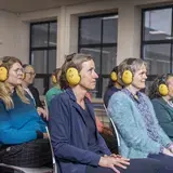 Een groep collega's met gele gehoorbeschermers luistert tijdens de cursus Ervaar Gehoorverlies..