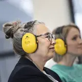 Een vrouw met een gele gehoorbeschermer kijkt naar voren, met een collega op de achtergrond.
