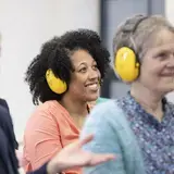 Een lachende vrouw met gele gehoorbeschermer, tussen collega's tijdens een workshop.