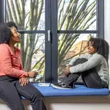 Een vrouw en kind zitten tegenover elkaar op een vensterbank met kaarten, in gesprek.