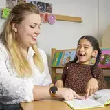 Een logopedist leest samen met een meisje in een boek. Ze wijzen allebei naar de tekst.