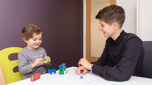 Een logopedist zit aan een tafel tegenover een peuter. Het kind speelt met speelgoedauto's en lacht. De logopedist kijkt toe en maakt aantekeningen.
