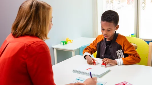 Een kind werkt met rode blokjes aan een taak. Tegenover hem zit een therapeut die aantekeningen maakt. Het kind is geconcentreerd bezig.