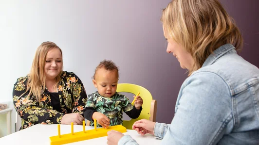 Een baby speelt met een geel speelgoedrek aan een tafel. Naast het kind zit de moeder. Tegenover hen zit een therapeut die het kind begeleidt.