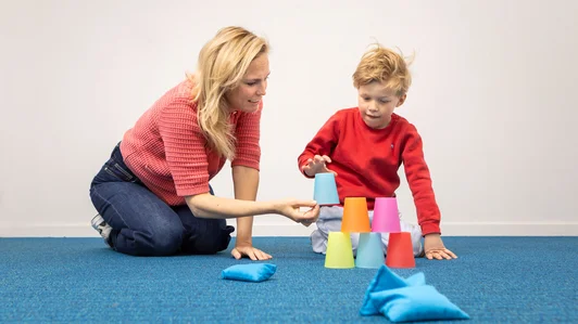 Een leerling bouwt een piramide van gekleurde bekers, met een begeleider ernaast.