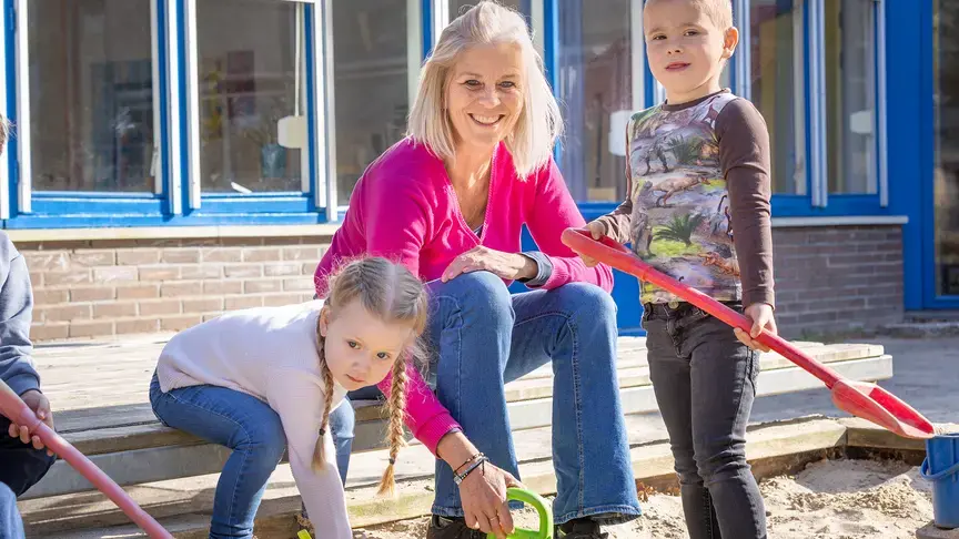 Leerkracht zit bij de zandbak terwijl twee kinderen spelen met scheppen.