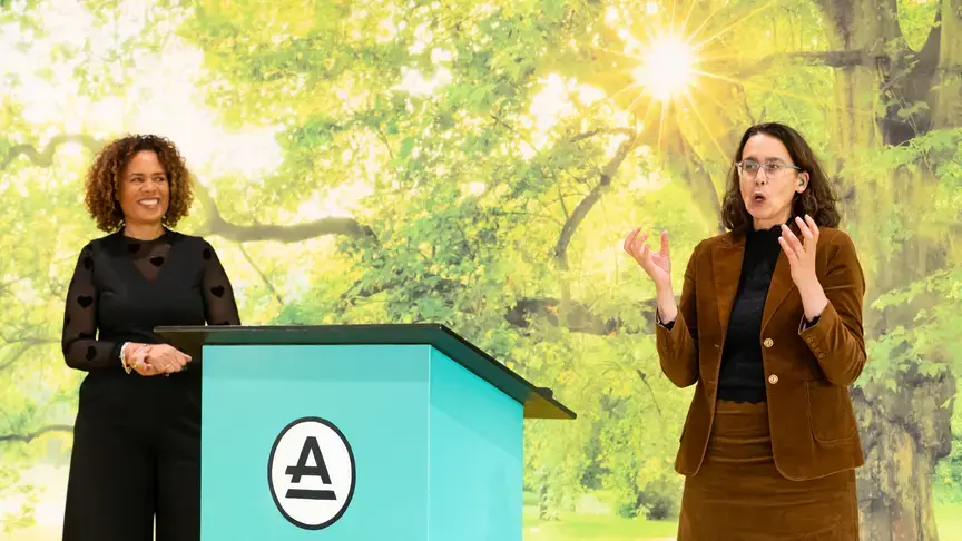 Twee vrouwen staan naast een spreekgestoelte met het Auris-logo, tijdens een presentatie.