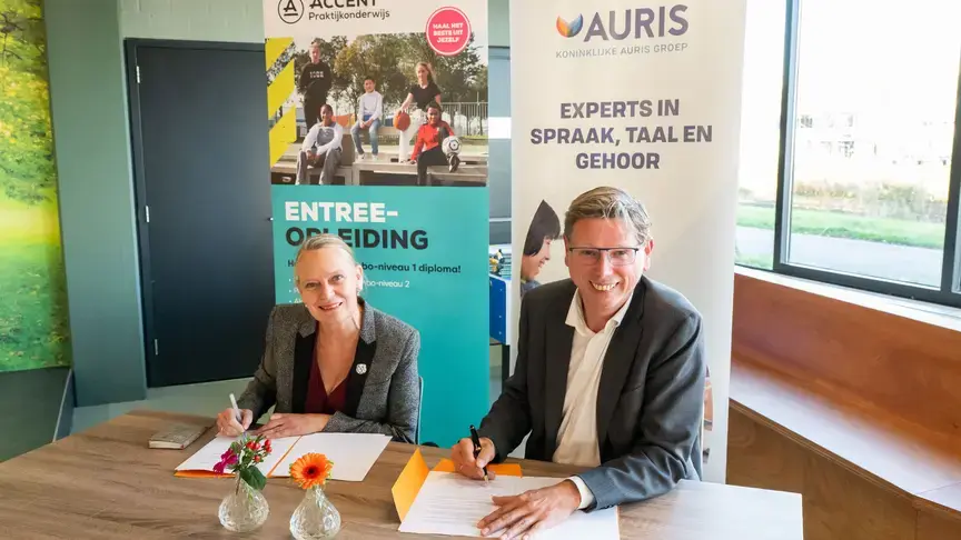 Twee mensen ondertekenen een overeenkomst aan een tafel, met banners van Auris en Accent op de achtergrond.