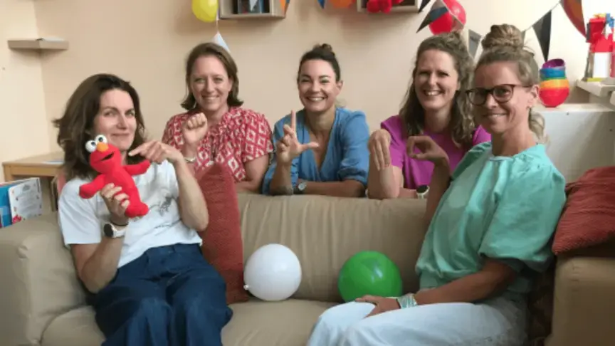 Vijf vrouwen zitten op een bank met een Elmo-knuffel en ballonnen, tijdens een opening.