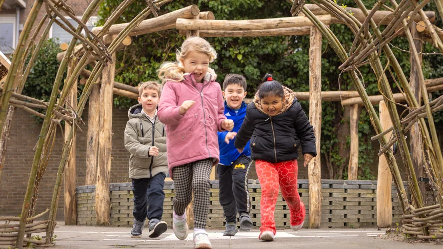 Vier leerlingen rennen lachend door een houten tunnel op het schoolplein.