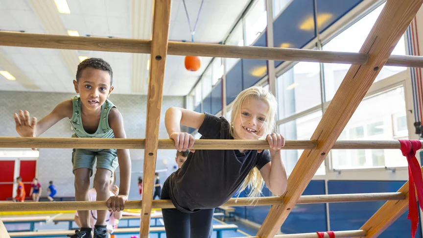 Twee leerlingen kijken lachend door de sporten van een klimrek in de gymzaal