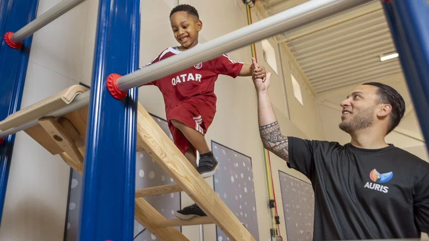 Een jongen loopt over een houten ladder in de gymzaal. De gymleraar helpt de jongen op de ladder.