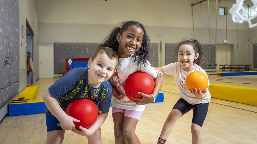 Drie kinderen met gymballen kijken lachend in de camera in een gymzaal.