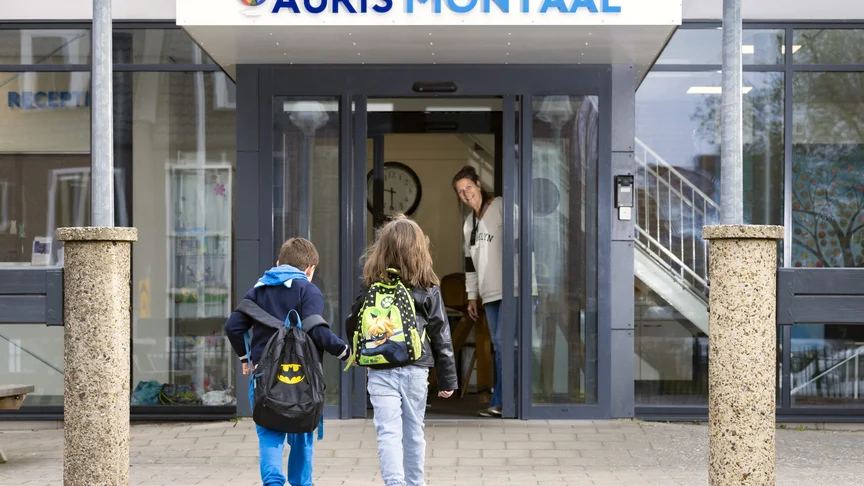 Twee kinderen met rugtas lopen naar binnen bij Auris Montaal, een begeleider staat in de deuropening