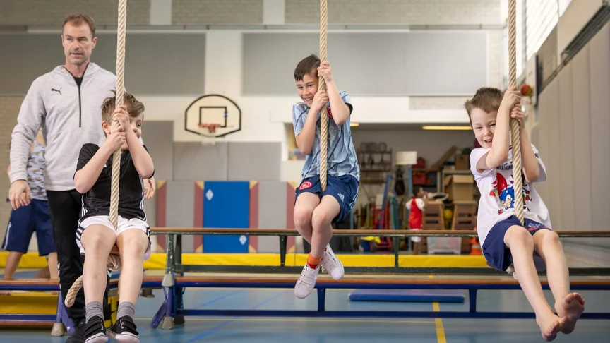 Een begeleider helpt 3 leerlingen bij het touwklimmen in de gymzaal. De leerlingen lachen en klimmen omhoog.