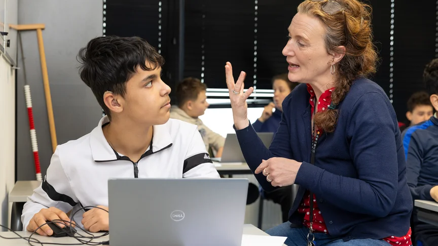 Een leraar met gekruiste vingers legt iets uit aan een jongen met een hoorbaar hoortoestel, die achter een laptop zit in de klas.