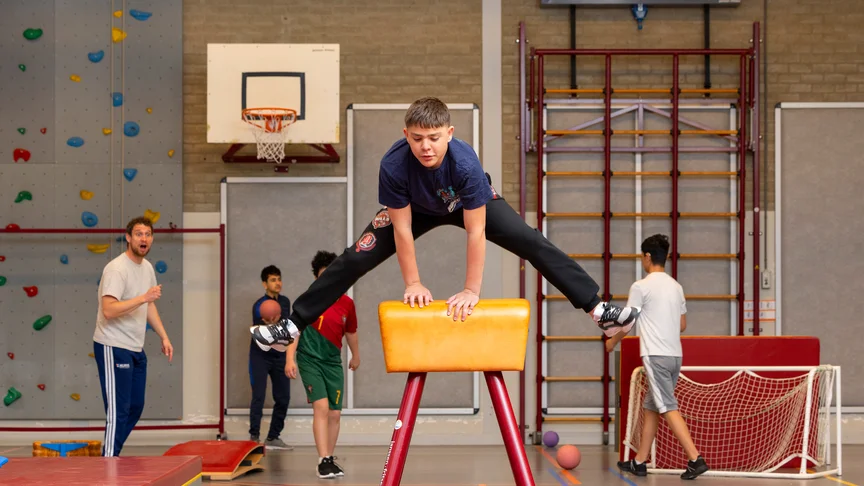Een jongen maakt een spreidsprong over de pegasus in de gymzaal, terwijl een leraar en klasgenoten op de achtergrond toekijken.