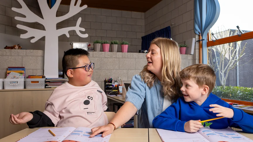 Een leerkracht praat met twee jongens aan tafel, met werkboekjes voor hen.