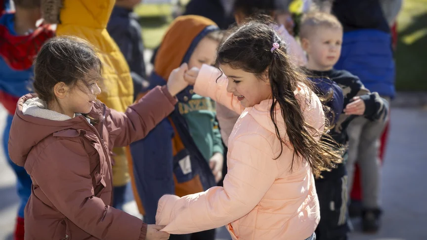 Twee meisjes stoten hun ellebogen tegen elkaar buiten, tussen andere kinderen.