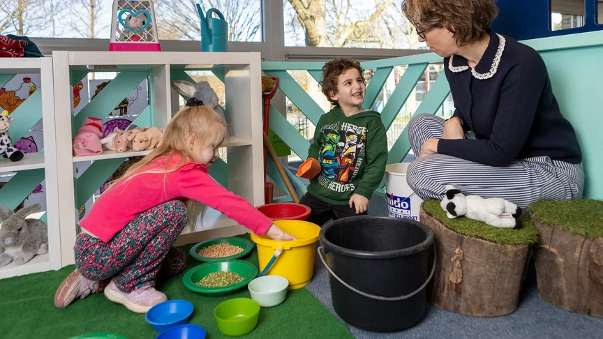 Twee leerlingen spelen met emmers en speelgoeddieren, terwijl een begeleider toekijkt.