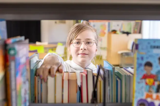 Meisje met bril kijkt door boekenrek in de schoolbibliotheek.