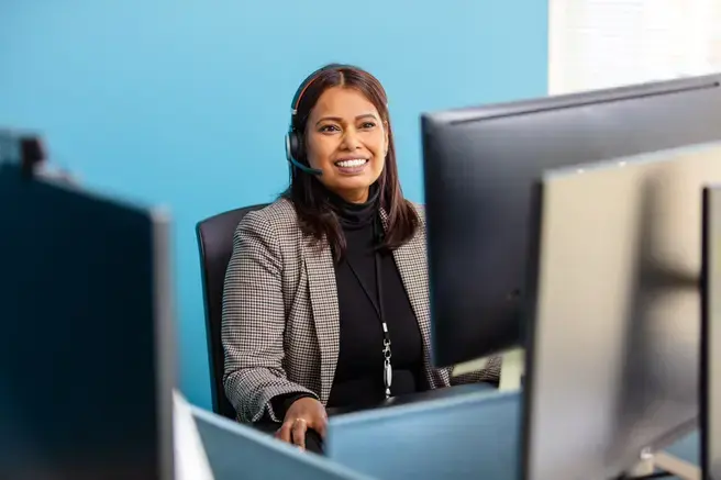 Een medewerker van het Auris Audiologisch Centrum zit aan haar bureau met een headset op. Ze glimlacht in de camera. Op de achtergrond is een blauwe muur zichtbaar.