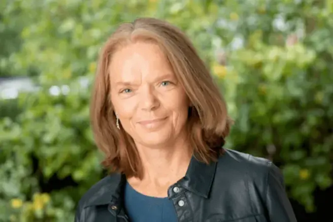 Vrouw met leren jasje aan kijkt in de camera. Op de achtergrond groene struiken.