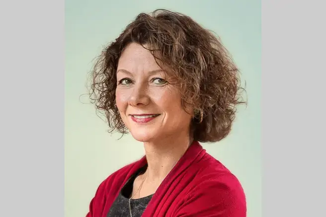 Vrouw met bruine krullen en rood jasje kijkt in de camera.
