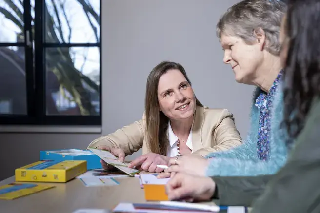 Een vrouw laat kaarten zien aan een vrouw aan tafel, met communicatiemateriaal ernaast.